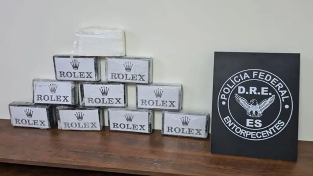Polícia Federal apreende 10 tabletes de cocaína tipo “Rolex” vindas da Argentina na Serra Sobre a mesa da Polícia Federal dez tabletes de cocaína embrulhadas com a marca Rolex, empilhadas e do lado direito uma placa de divulgação da Polícia Federal