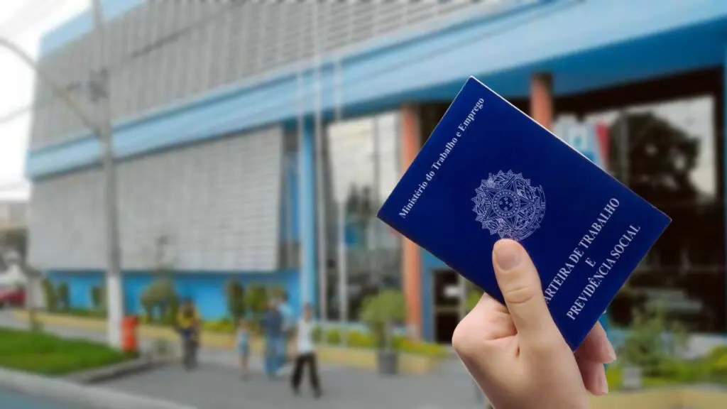 Sine de Vitória conta com vagas abertas para trabalhar em nova unidade de empresa de telecomunicações Arte com uma mão segurando uma carteira de trabalho e ao fundo desfocado a sede do Sine Vitoria