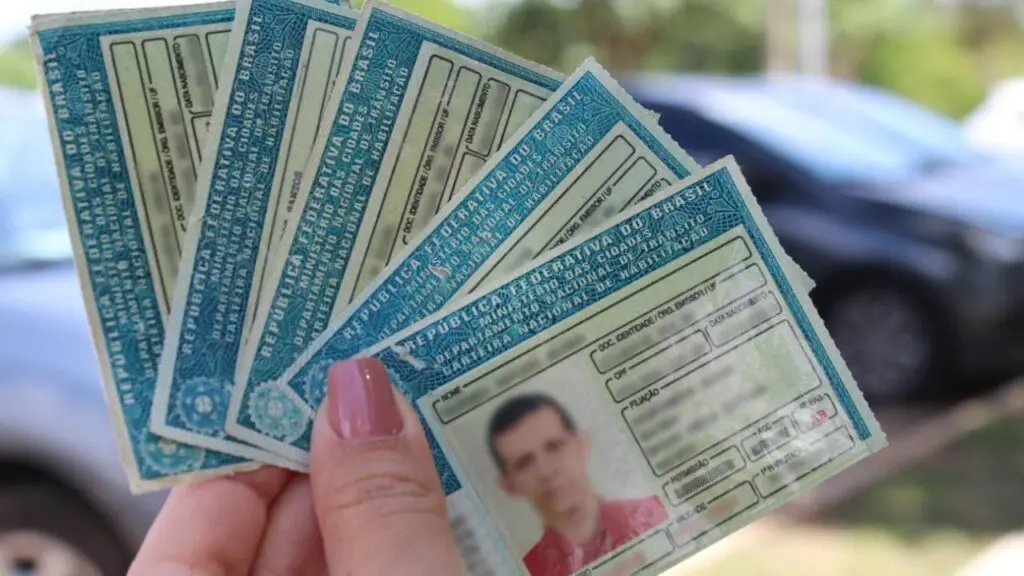 Imagem de uma mão segurando 5 Carteiras de motorista, e ao fundo desfocado carros estacionados