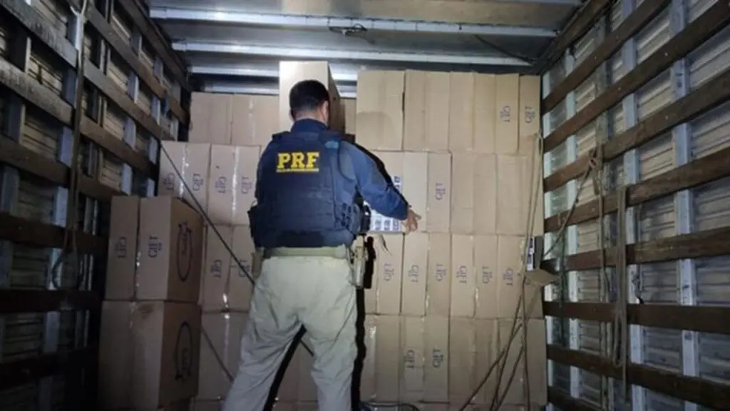 PRF flagra caminhão com carregamento ilegal de 100 mil maços de cigarros do Paraguai no ES Foto de um agente da Polícia Federal dentro de um baú de caminhão, em frente a pilhas de caixas de cigarros que estava sendo contrabandeando