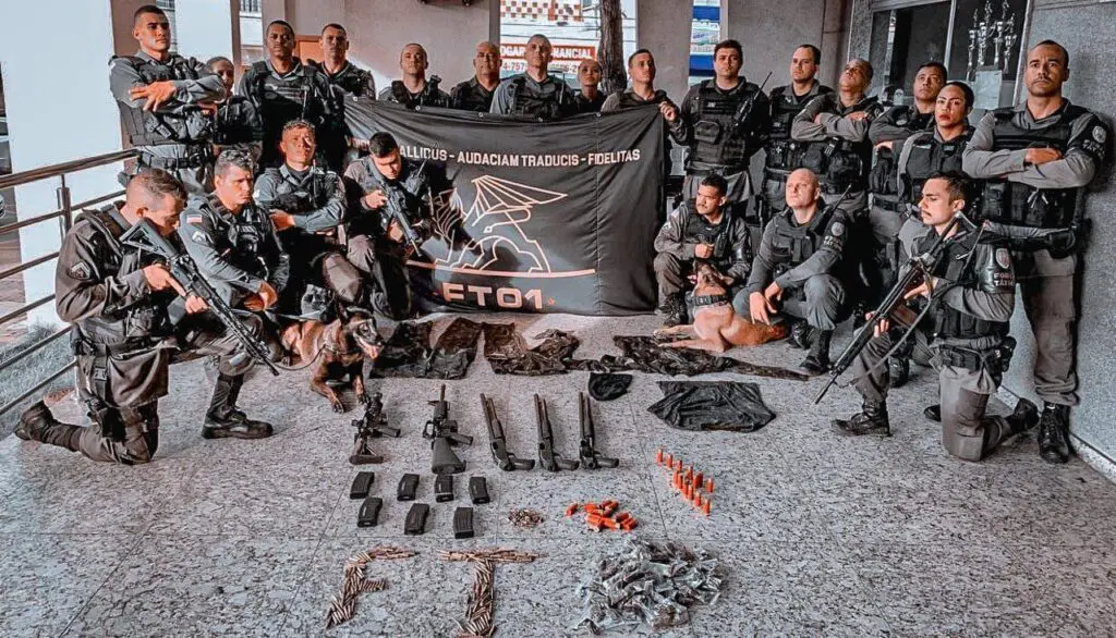 22 Policiais da Força Tática e um cão farejador posando em uma sala com o material apreendido três espingardas calibre 12, dois fuzis calibre .556, sete carregadores de fuzil calibre .556, 310 munições calibre .556, 33 munições calibre 9mm, 28 munições calibre .12, 131 buchas de maconha além de algumas roupas camufladas durante a operação