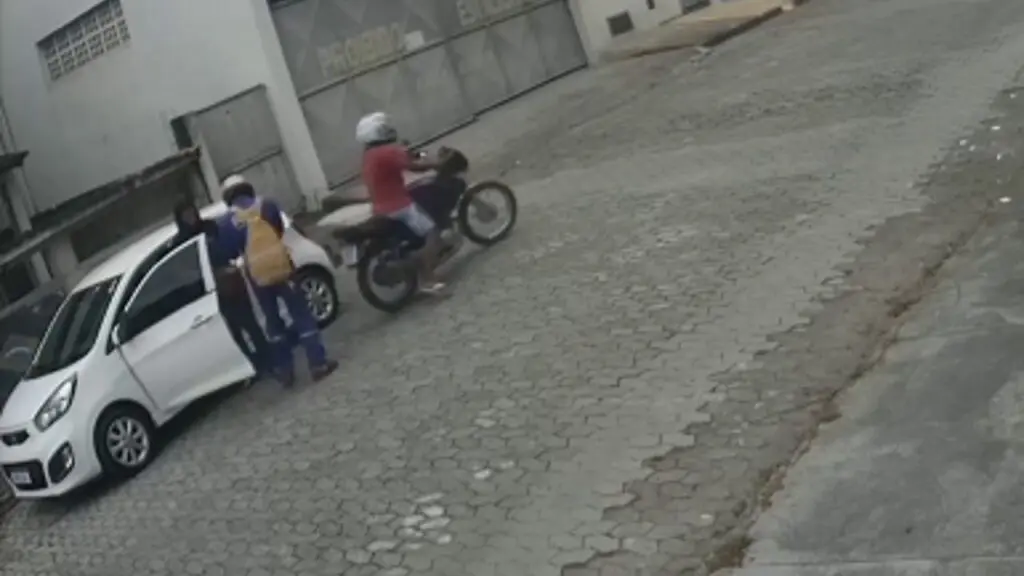 Vídeo: câmera de segurança flagra homem de uniforme assaltando mulher na Grande Vitória imagem de um veículo branco parado em uma rua de paralelepípedo e uma moto com dois homens assaltando a motorista que está parada em pé ao lado do veiculo