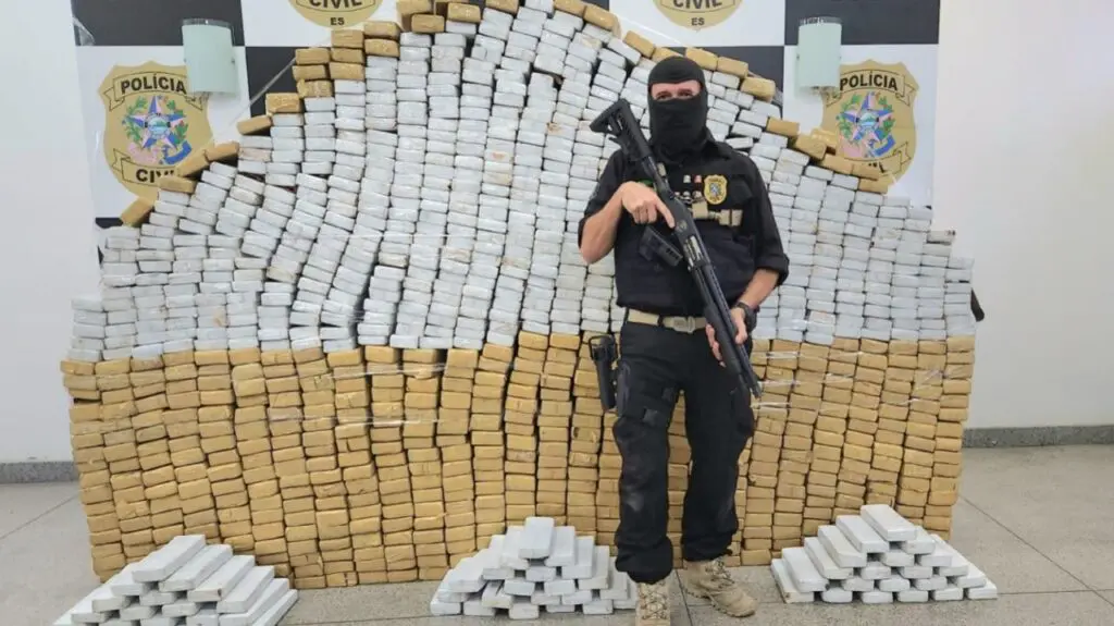 Operação da Polícia Civil resulta em mega apreensão de uma tonelada de maconha em Guarapari Um paredão de com centenas de tabletes de drogas e um Agente da Polícia Militar armado e de capuz posando para a foto de divulgação.