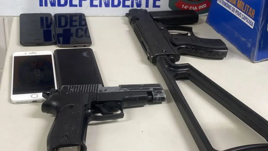 Polícia Militar chega antes e evita arrastão dentro de ônibus do Transcol na Serra Foto de materiais apreendidos durante uma ocorrência em um ônibus do sistema Transcol, sobre uma mesa branca uma réplica de uma pistola com o pente alongado e um simulacro de um fuzil além de 4 celulares