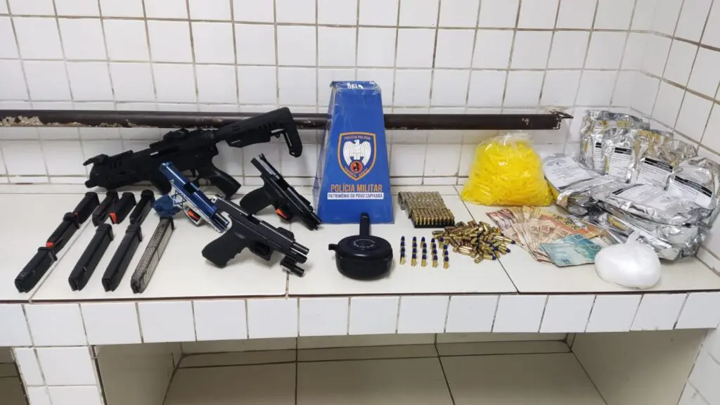 Foto de materiais apreendidos pela Polícia Militar, sobre uma bancada na Delegacia Regional situada na Serra, três pistolas de diferentes marcas e modelos, todas calibre 9mm, acessórios que aumentam o potencial ofensivo das armas, treze carregadores de pistola, 242 munições de calibre 9mm, sete munições de calibre .40, além de 500 gramas de cocaína e substâncias comumente usadas para refino de cocaína além de R$ 1.879,00 reais em espécie.