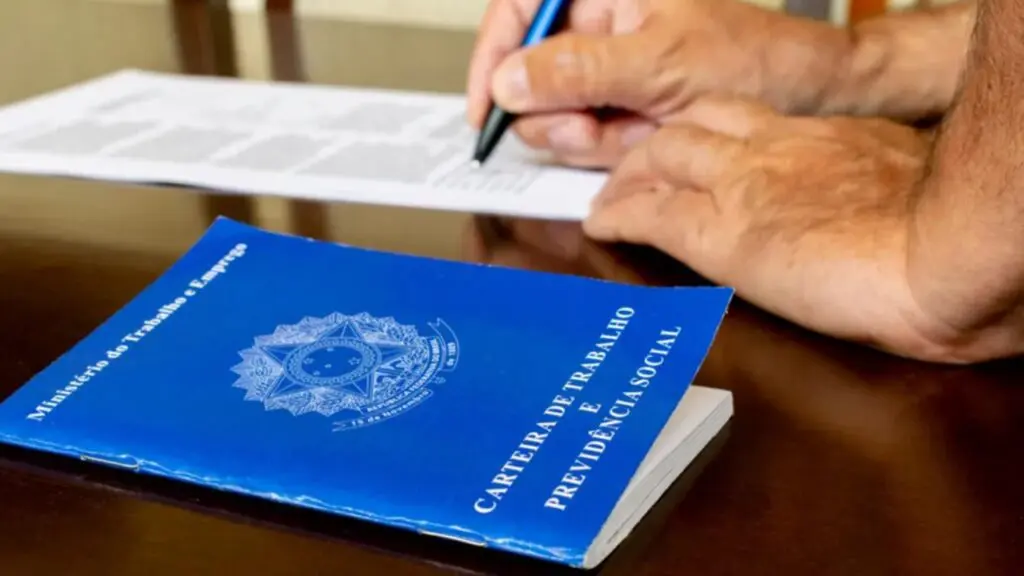 Foto de um homem assinando um contrato de trabalho, com a carteira de trabalho ao lado.