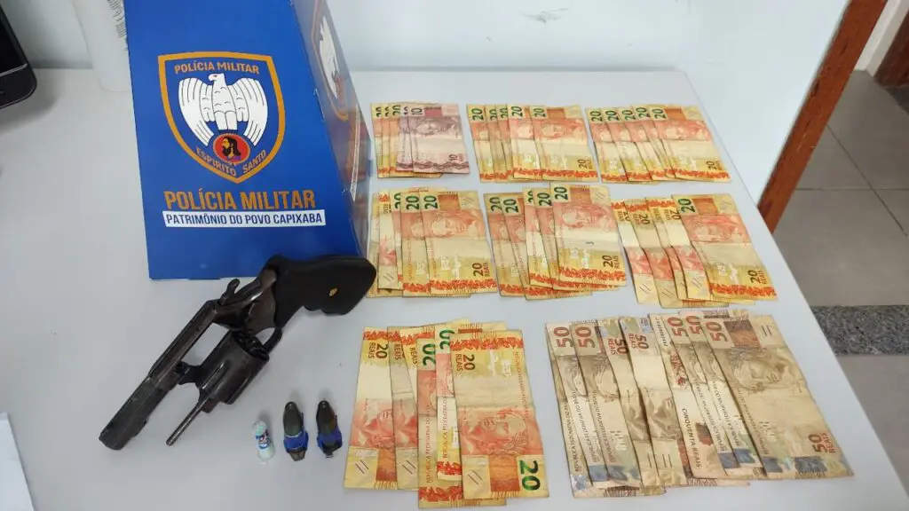 Foto de materiais apreendidos durante patrulhamento da PM, sobre uma mesa branca, um revólver calibre .38, 3 pinos de cocaína, além de dezenas de notas contabilizando 1.130 reais
