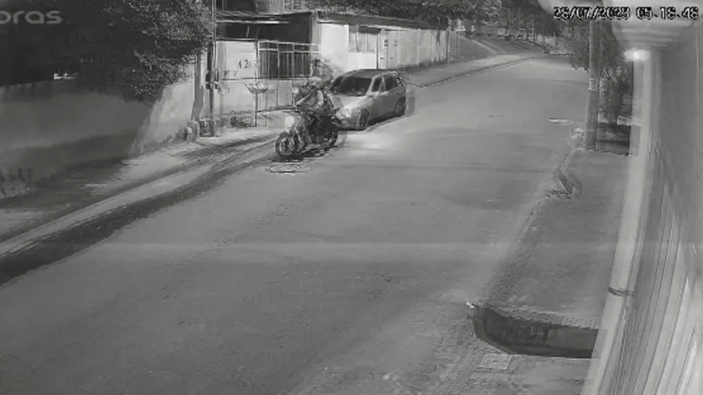 Vídeo: câmera flagra dupla furtando moto durante madrugada em Jardim Bela Vista Foto preto e branco de uma rua residencial, com um automóvel estacionado, e um ladrão pilotando uma moto, parado esperando seu comparsa realizar o furto de outra motociclieta.