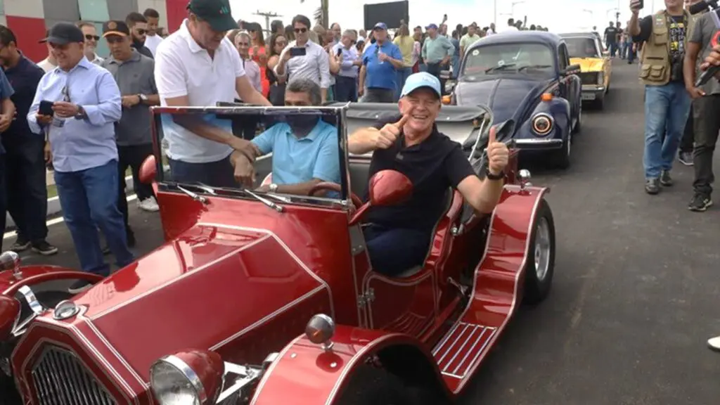 Renato Casagrande dentro de um veículo conversível, modelo bem antigo "Ximbica" fazendo sinal de positivo com a mão, na inauguração de uma importante obra de mobilidade, ao lado dele o Prefeito da Serra sendo ajudado por uma pessoa a entrar no veículo.