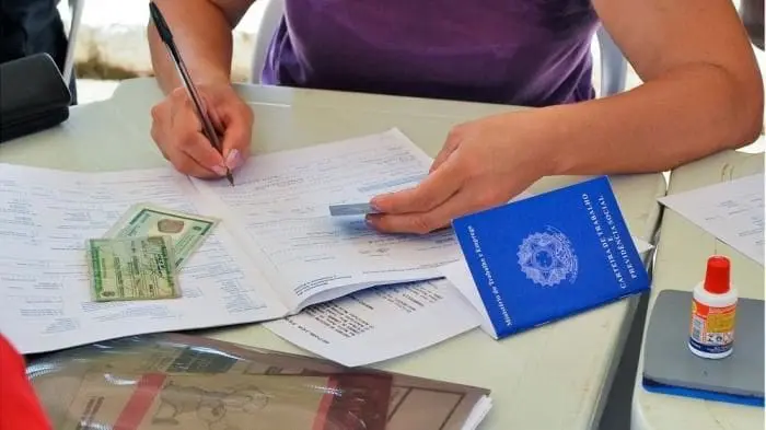 Uma mesa com alguns documentos, e uma atendente preenchendo uma ficha para o candidato poder concorrer a uma vaga de emprego