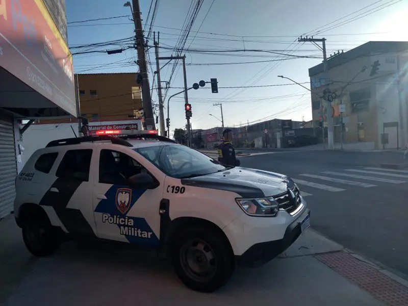 Uma viatura da Polícia Militar estacionada em uma via, publica com um militar ao lado.
