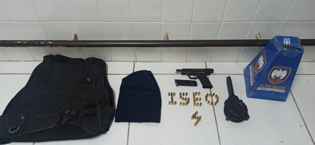 Polícia Militar apresentando o material apreendido, sobre um banco uma pistola calibre 9mm, um carregador com capacidade para 17 munições calibre 9mm, um carregador tipo caracol com capacidade para 50 munições calibre 9mm, carregado com 36 munições, totalizando 53 munições intactas, uma toca ninja e uma capa de colete.
