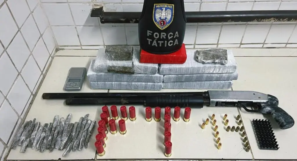 Materiais apreendidos no bairro das Laranjeiras, pelas equipes da Força Tática da Polícia Militar, sobre uma mesa uma escopeta calibre 12, 07 tabletes de maconha, 03 pedaços de maconha, 17 tiras de maconha, 20 munições de calibre 12, 10 munições 9mm, 10 munições .380, além de uma balança de precisão pequena.