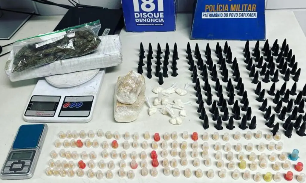 Materiais apreendidos no bairro de Feu Rosa, pelas equipes da Ronda Ostensivas da Polícia Militar, sobre uma mesa um tablete de maconha e uma sacola com restos de maconha totalizando aproximadamente 960g, 123 pinos de crack, 146 pinos de cocaína, duas pedras grandes de crack, além de 02 balanças de precisão.
