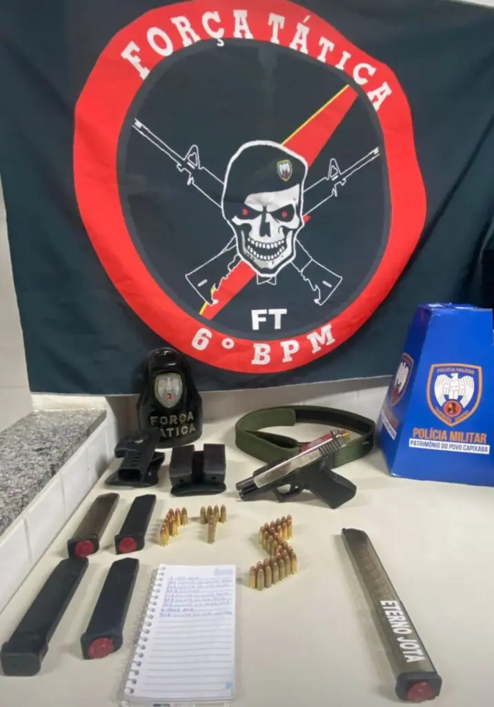 Materiais apreendidos sobre uma mesa durante a prisão do traficante, uma pistola calibre 9mm com carregador alongado, um cinto tático contendo dois carregadores sobressalentes, um coldre de arma de fogo e uma bolsa com mais dois carregadores, totalizando 46 munições de calibre 9mm, um caderno de contabilidade do tráfico de drogas e um celular. E no fundo na parede a bandeira da Força Tatica do 6º Batalhão da PM, com uma caveira de boina e dois fuzis cruzados.