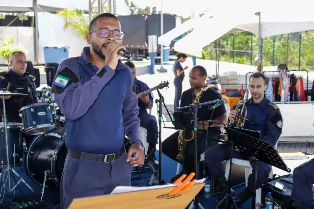 Musicos da Banda da Guarda da Serra se apresentando em um evento. O vocalista em pé cantando e outros músicos em volta dele tocando Baixo, guitarra, instrumentos de sopro e bateria.