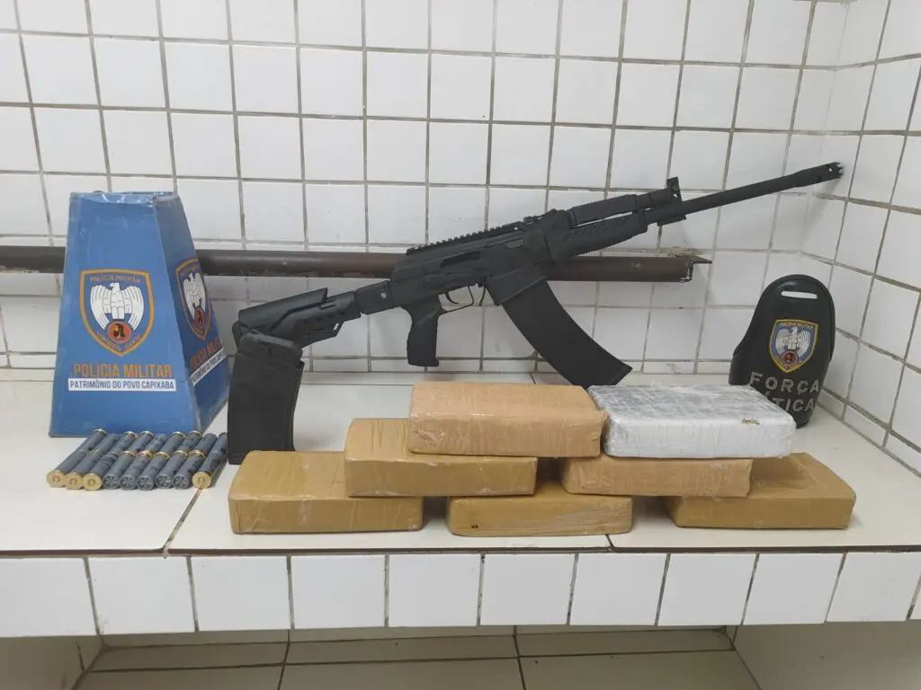 Materiais apreendidos pela equipe da Força Tática sobre uma bancada em uma sala da delegacia 7 tabletes de crack, 18 munições intactas de cal .12, espingarda cal. 12, carregador sobressalente alongado cal. 12 com capacidade para 11 munições.