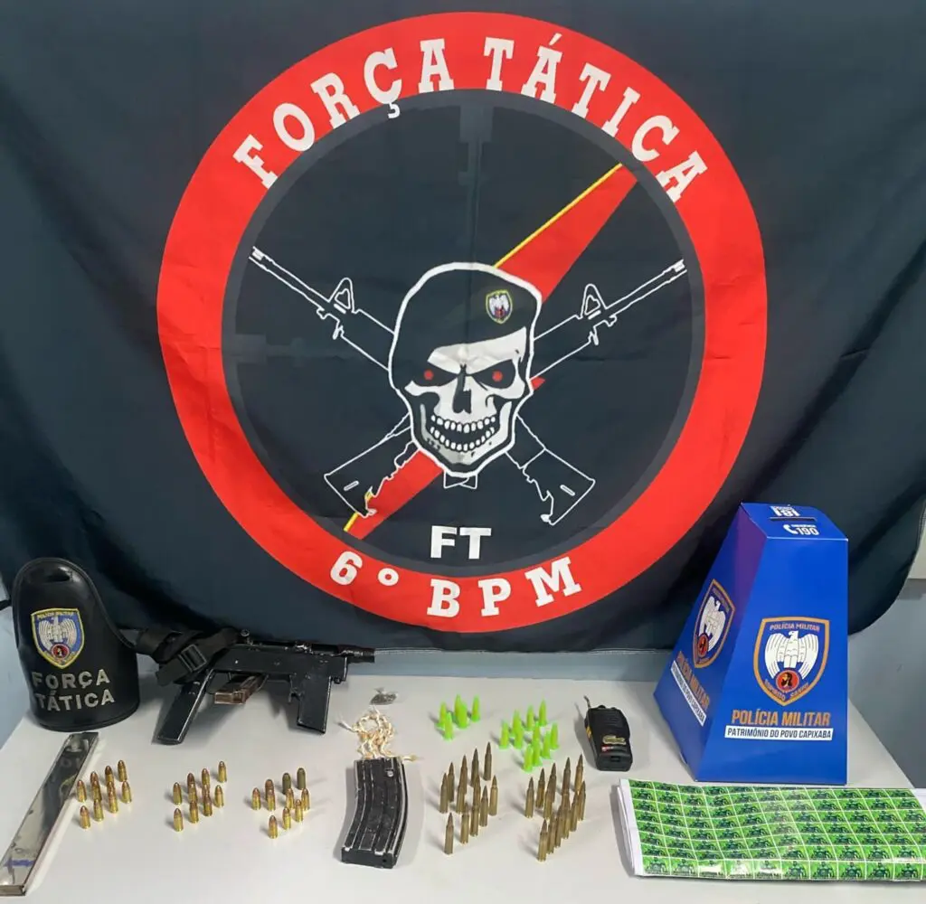 Materiais apreendidos pela Força Tática, sobre uma mesa uma metralhadora de fabricação caseira, dois carregadores, 55 munições de diferentes calibres, uma bucha de maconha, 30 pedras de crack, 15 pinos de cocaína além de um rádio comunicador.
