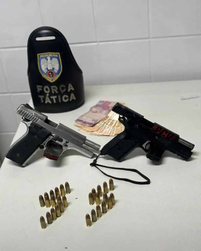 Sobre uma mesa armamentos apreendidos pela Força Tática: duas pistolas apoiadas em seus carregadores, uma pistola calibre .45, e outra calibre 9mm, 28 munições calibre 9mm, além de 655 reais em dinheiro. 