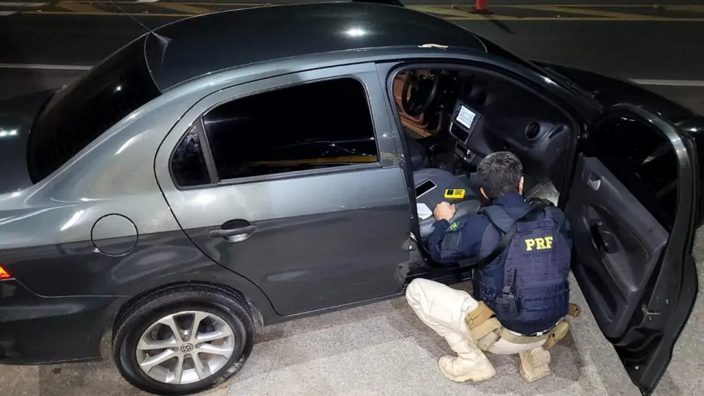 Um agente da Polícia Rodoviária Federal, abaixado ao lado de um carro com uma das portas abertas, vistoriando o veículo recuperado, durante uma abordagem.