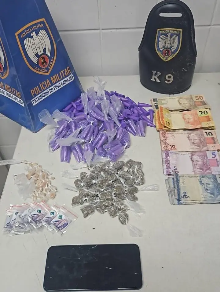 Materiais apreendidos pelas equipes da Força Tática, sobre uma mesa uma braçadeira de couro da Equipe K9, um material de divulgação de papelão da Polícia Civil em forma de pirâmide, 221 pinos de cocaína, 35 pedras de crack, 13 buchas de haxixe, 50 buchas de maconha, R$ 513 em espécie, além de um celular.