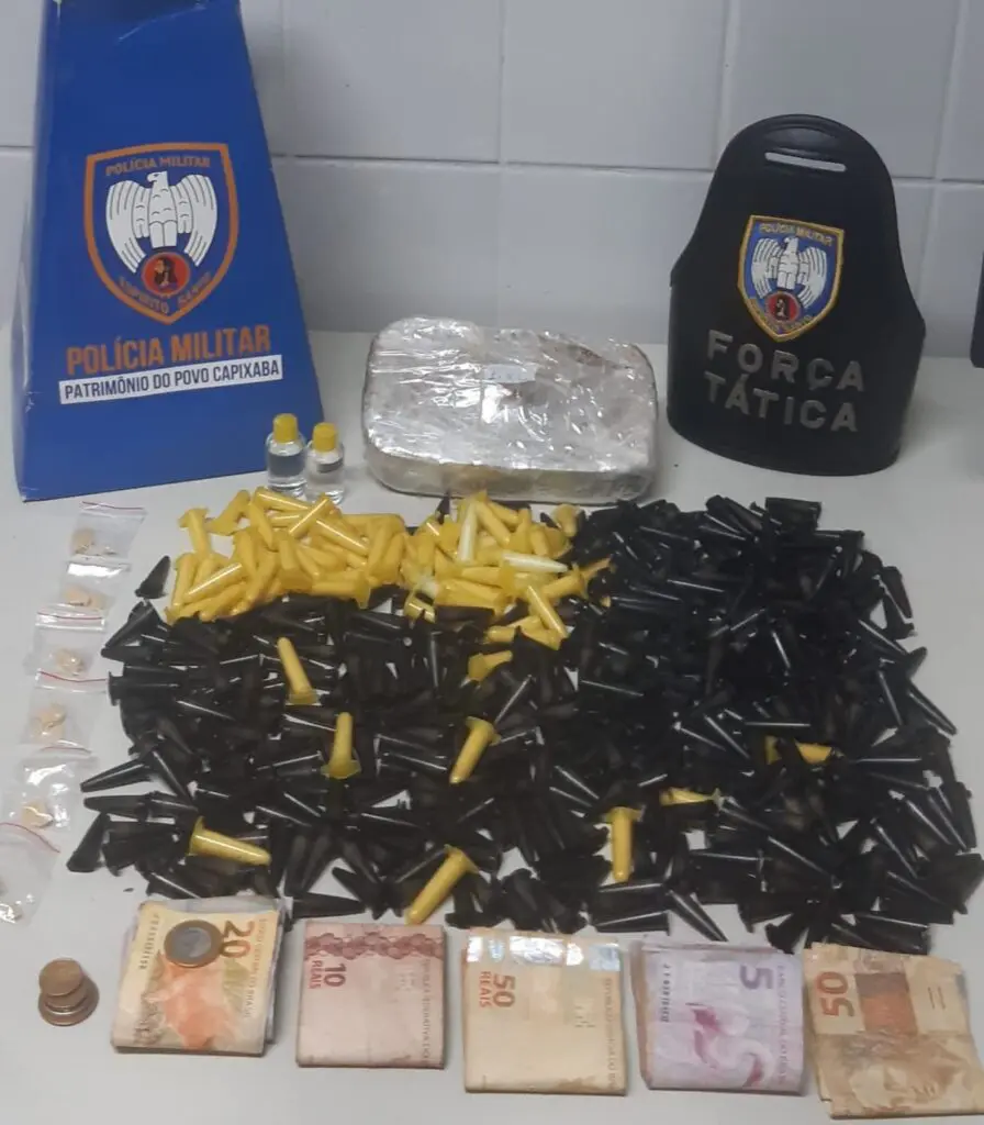 Materiais apreendidos pela equipe da Força Tática sobre uma mesa, uma braçadeira da Força Tática, um cone de papelão com o emblema da Polícia Militar do Espírito Santo, várias cedulas de dinheiro, 447 pinos de cocaína, 6 pedras de crack, 2 unidades de Loló além de um tablete de crack de aproximadamente 1 kg.