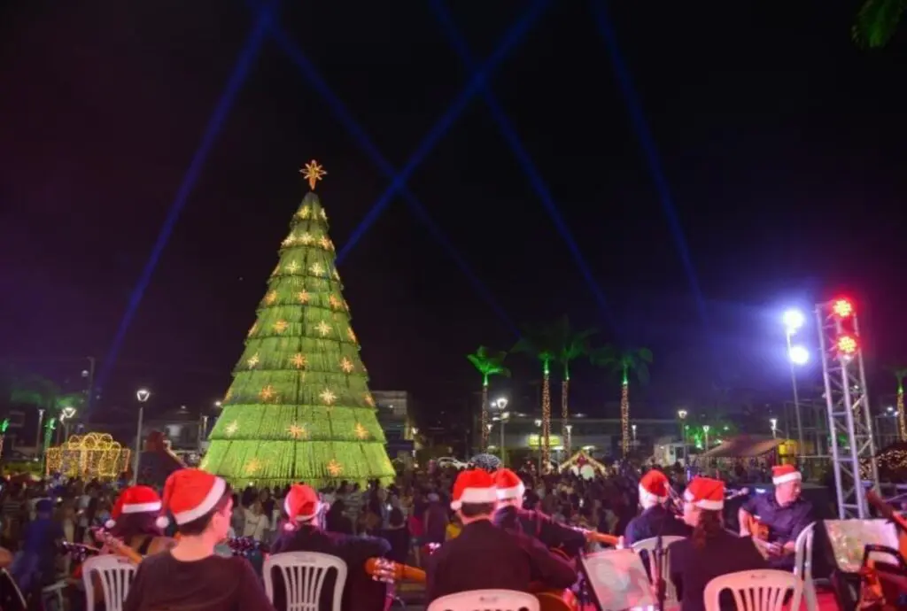 Imagem de um parque com decorações natalinas e uma árvore de Natal ao centro, com as luzes acesas, em volta diversos músicos se apresentando com chapeis de Natal.