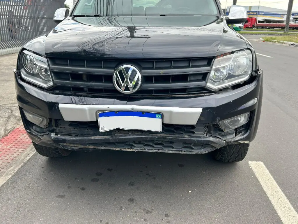 Um veículo modelo picape Amarok, com o para-choque amassado após um motociclista se chocar contra o veículo.
