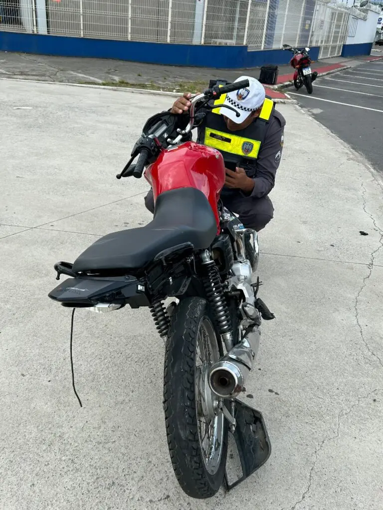 Uma motocicleta, após um acidente com vários danos em sua estrutura, com um policial militar abaixado próximo a ela realizando a consulta do chassis.