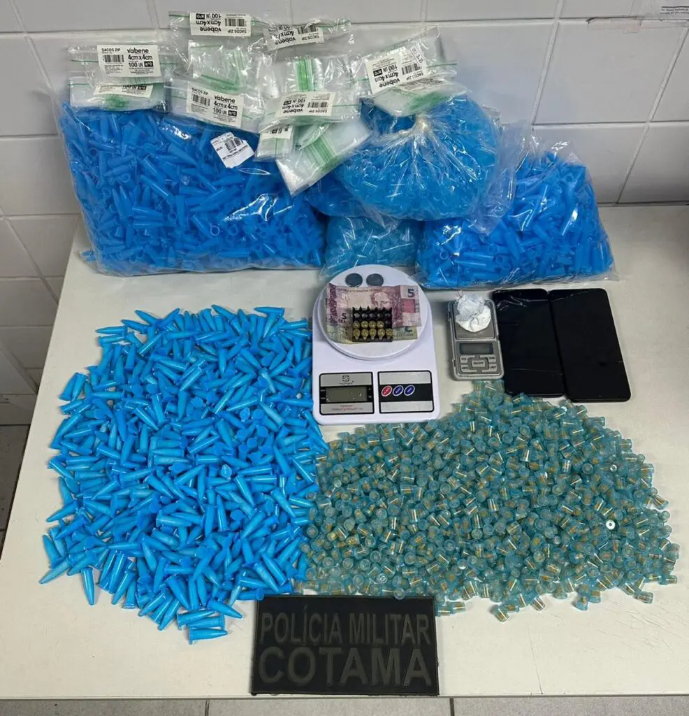 Sobre uma mesa uma apreensão da Polícia Militar, com milhares de pinos com cocaína e crack, 2 balanças de precisão, pinos vazios, dinheiro em espécie e um material de divulgação da equipe do Comando de Operações Táticas em Mata Atlântica (COTAMA).