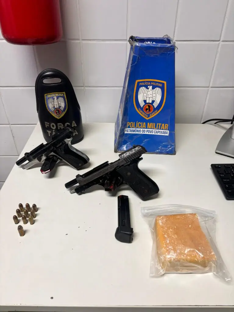 Imagem de uma mesa com materiais apreendidos, durante uma prisão de um homem procurado pelo crime de homicídio, sobre uma mesa duas pistolas calibre .380, três carregadores, dez munições e uma pedra grande de crack.
