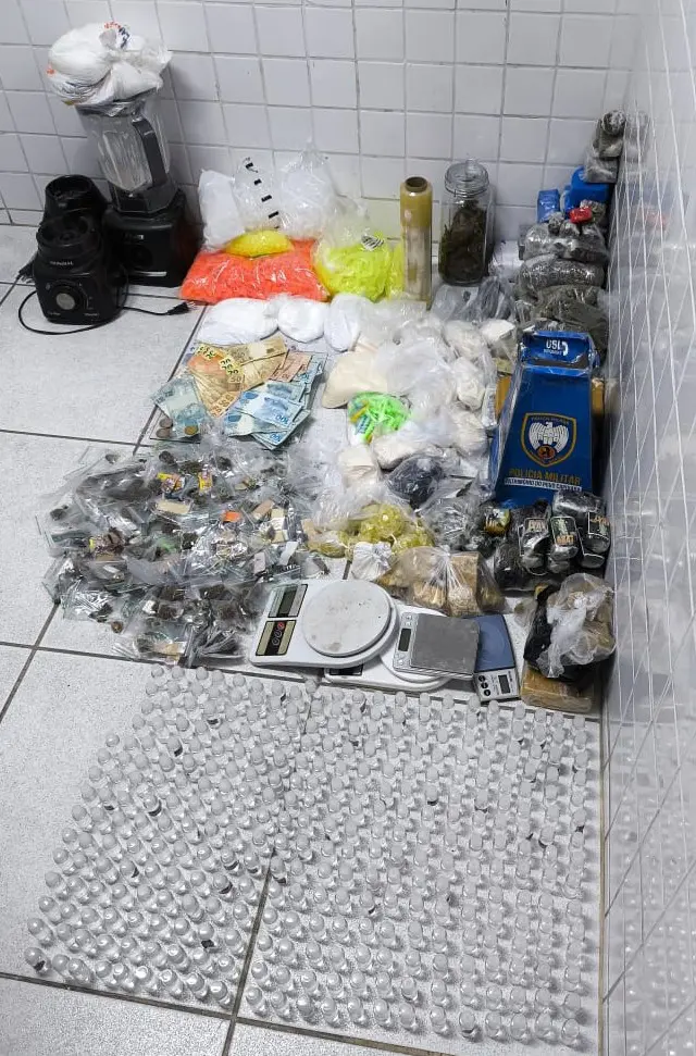 Sobre o chão de uma delegacia diversos materiais apreendidos durante uma ação da Polícia Militar: Tabletes, pedaços e sacos de maconha, pinos de cocaína, frascos de lolo, matérias usados para preparo e embrulho de drogas e dinheiro.