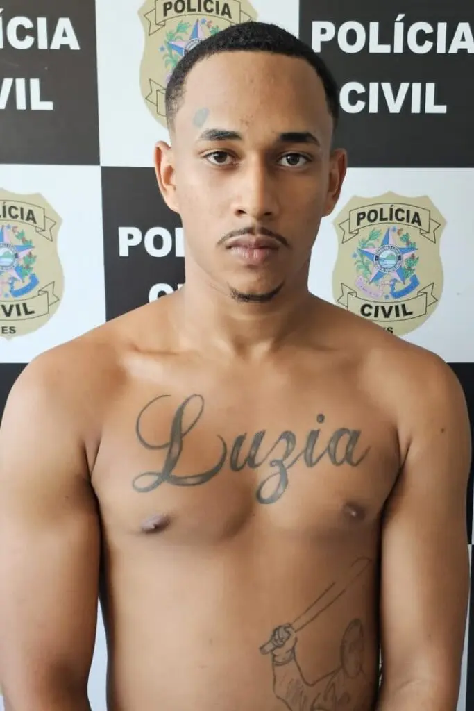 Imagem de um homem (Alan Carlos de Souza Elias, de 18 anos), parado em frente a uma parede com informações de divulgação da Polícia Civil/DHPP.