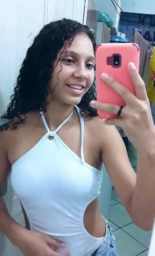 Uma jovem (Fernanda Prates Santos Bernardo, de 19 anos) segurando um celular próximo ao rosto fazendo uma selfie.