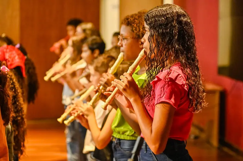 Cerca de 8 alunos tocando flauta doce, em um teatro.