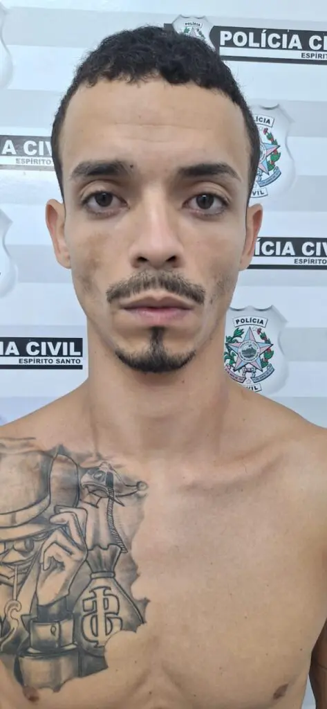 Imagem de um homem de 29 anos presos por cometer diversos crimes, (Tiago de Jesus Fernandes), sem camisa parado em frente a uma parede com imagens de divulgação da Policia Civil.