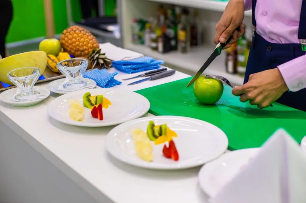 Imagem de uma competição de culinária com um homem cortando uma maça verde para usar em dois pratos que estão sobre a mesa.