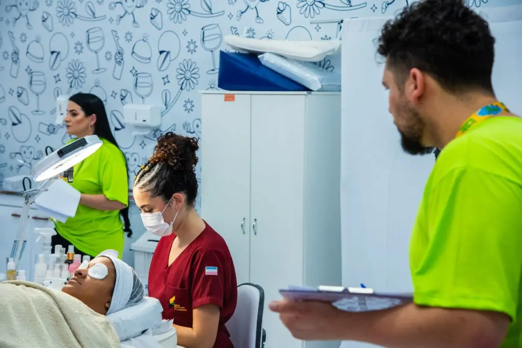 Uma jovem participando de uma competição de estética, com uma pessoa deitada em uma maca e enquanto ela realiza procedimentos de beleza na pessoas, dois juízes estão ao seu lado avaliando ela.