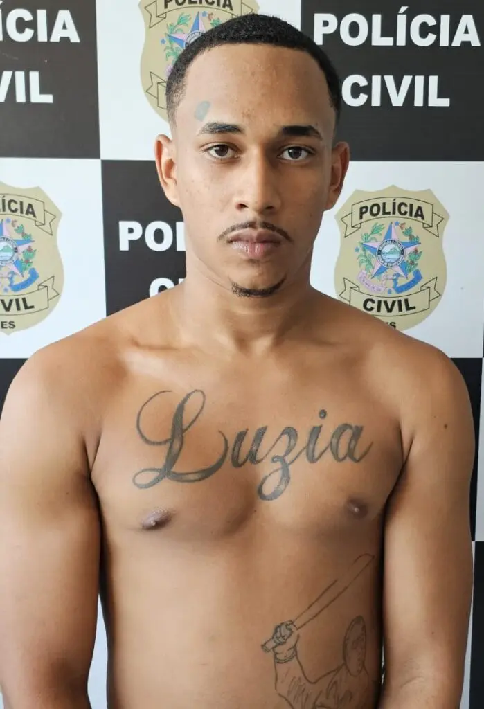 Imagem de um sujeito (Alan Carlos de Souza Elias, vulgo “Sombra”, de 18 anos) em frente a um banner de divulgação de prisões da Polícia Civil.
