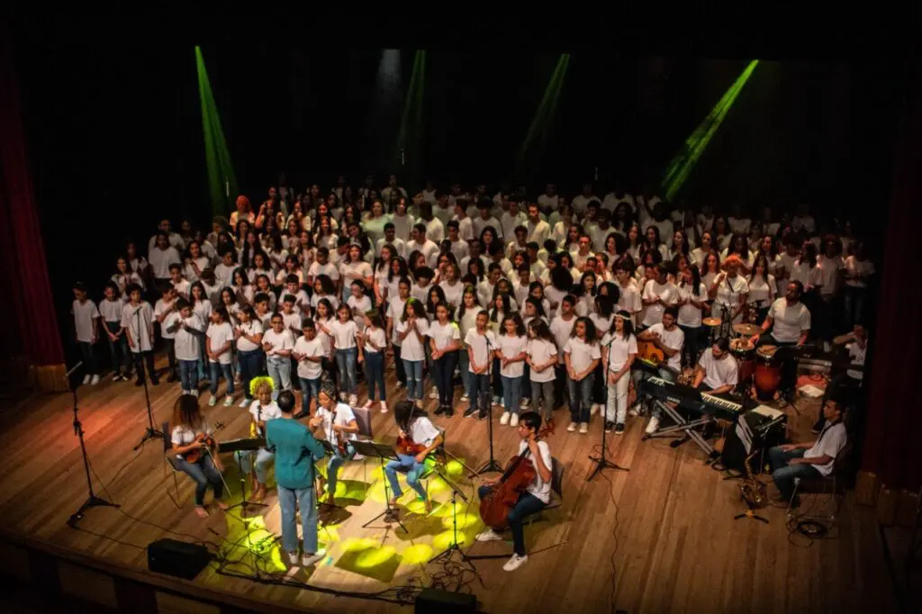 Imagem de um maestro regendo um coral formado por um grupo de crianças de idades entre 7 e 11 anos em um palco de um teatro.