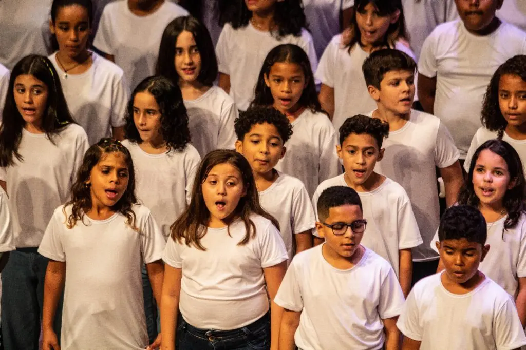 Imagem de um grupo de crianças de idades entre 7 e 11 anos reunidas cantando durante uma aula de canto.