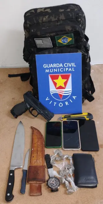 Imagem dos materiais apreendidos durante uma ação da Guarda de Vitória onde foram recolhidos: duas facas uma grande e outra pequena, 16 buchas de maconha, um estilete, três celulares, uma carteira, uma mochila, além de uma réplica de uma pistola. Todo o material está sobre o chão com uma placa de identificação da Guarda Civil Municipal de Vitória.