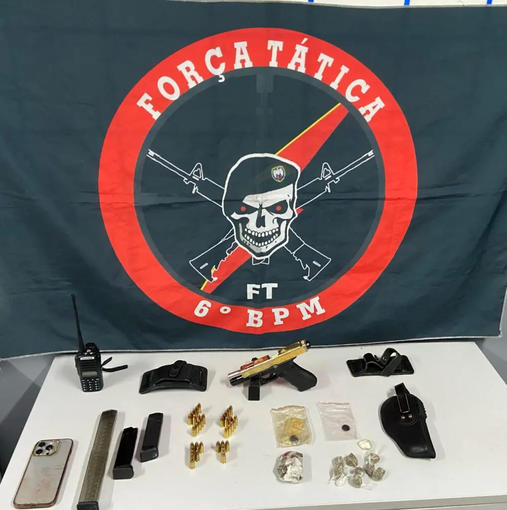 Uma sala com uma bandeira da Força Tática do 6º Batalhão, após uma ocorrência envolvendo traficantes, sobre uma mesa uma pistola, três carregadores, munições e drogas. 