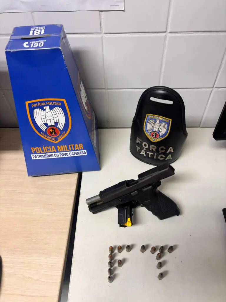 Imagem de uma pistola, 15 munições, um totem de papelão de divulgação da PMES e uma braçadeira de identificação da Força Tática sobre uma mesa de uma delegacia.
