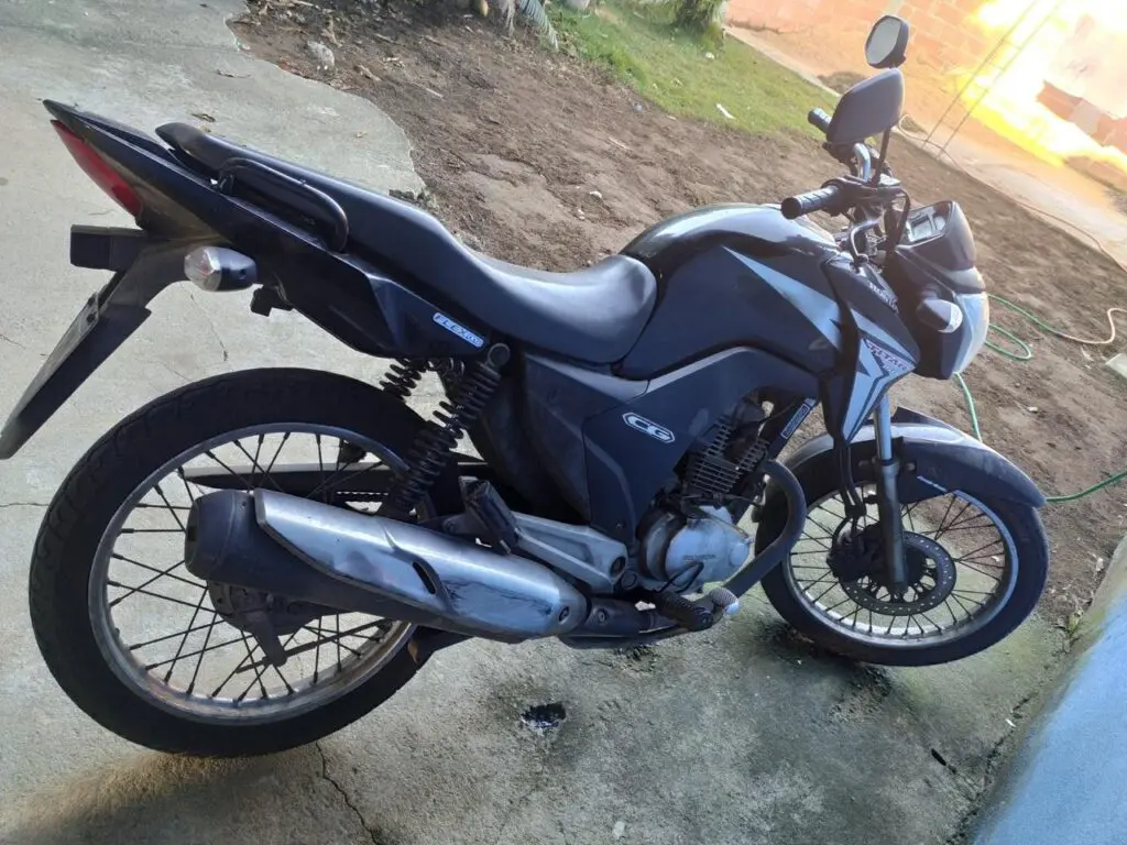 Uma motocicleta modelo Honda CG 150 Titan, estacionada em um quintal de uma residência.