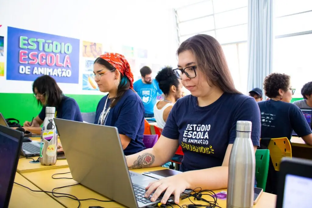 Uma sala de aula com cerca de 7 jovens sentado em frente a computadores, e no fundo da sala um professor explicando algo sobre animação, durante uma aula no Estúdio Escola de Animação.