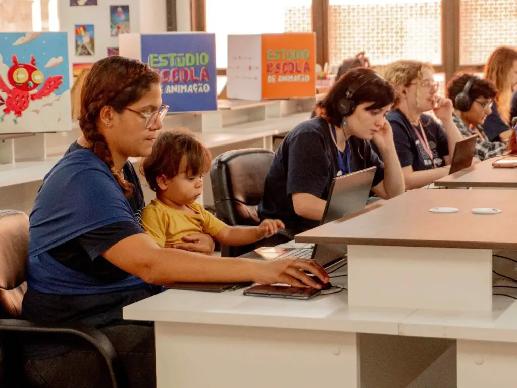 Uma sala de aula com 5 jovens sentado em frente a computadores durante uma aula de animação. Um dos alunos está junto com uma criança no colo.