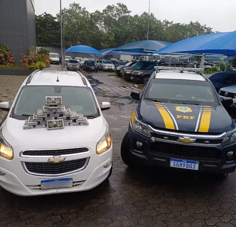 Em um patio onde há diversos veículos estacionado, em destaque um veículo modelo Chevrolet Spin branco com 27 tabletes de cocaína expostos empilhados sobre o seu capô e ao lado uma viatura da Polícia Rodoviária Federal.