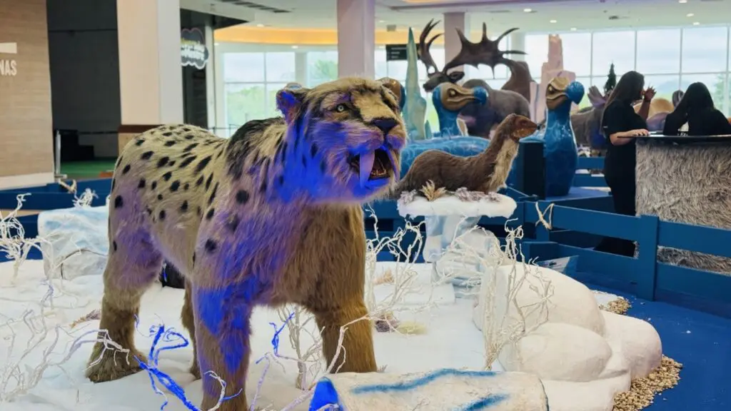 Imagem da Exposição “Gigantes da Era do Gelo”, no saguão de um shopping, com bonecos gigantes de um tigre dentes de sabre e outros animais pré-históricos, no fundo.
