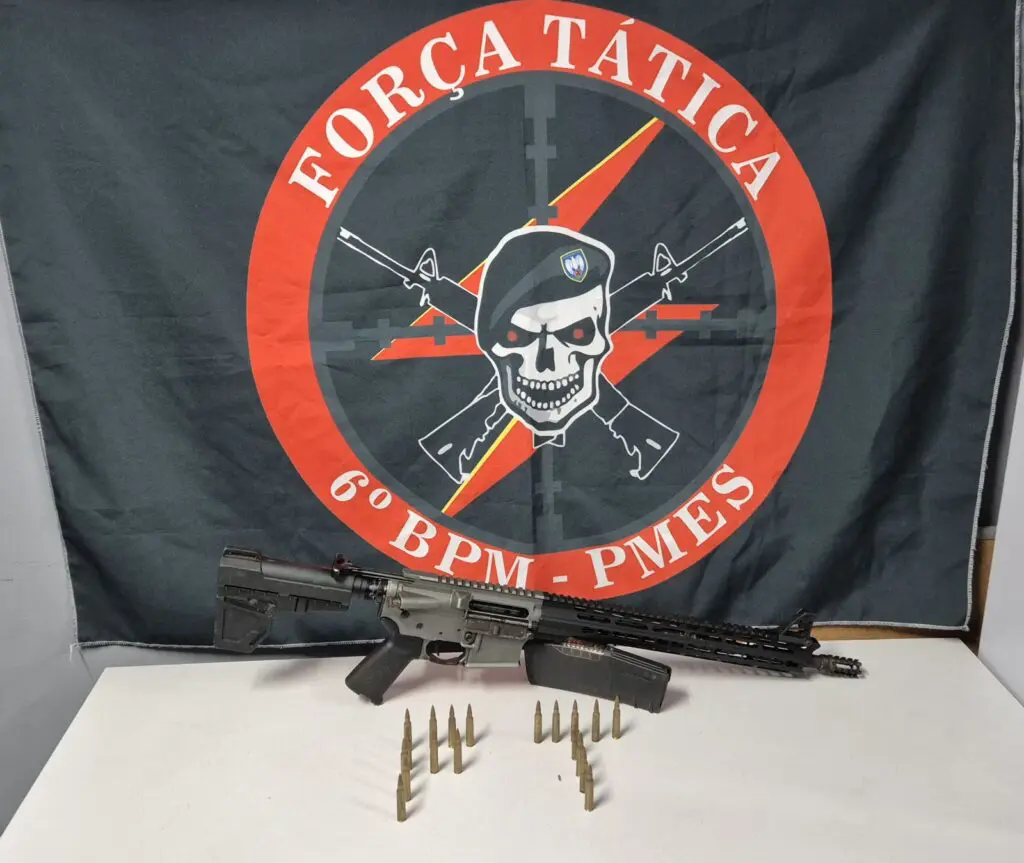 Sobre uma mesa um fuzil AR-15 calibre 5,56, um carregador com capacidade para 30 munições, e 19 munições. pendurada sobre a mesa uma bandeira da Força Tática do 6º Batalhão. (Uma caveira de boina e dois fuzis cruzados)